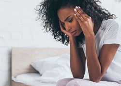 A young woman sitting on the side of her bed and holding her head because of an intense headache