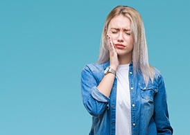A young woman wearing a denim top holding her cheek and cringing in pain from a toothache