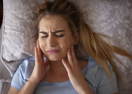 A young female lying in bed and holding both sides of her jaw in pain knowing she needs to see a dentist in Juno Beach for help