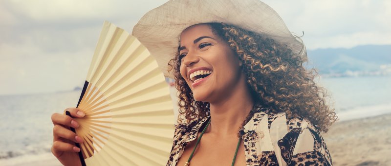 a young woman wearing a hat and carrying a hand fan while on the beach and showing off her beautiful smile